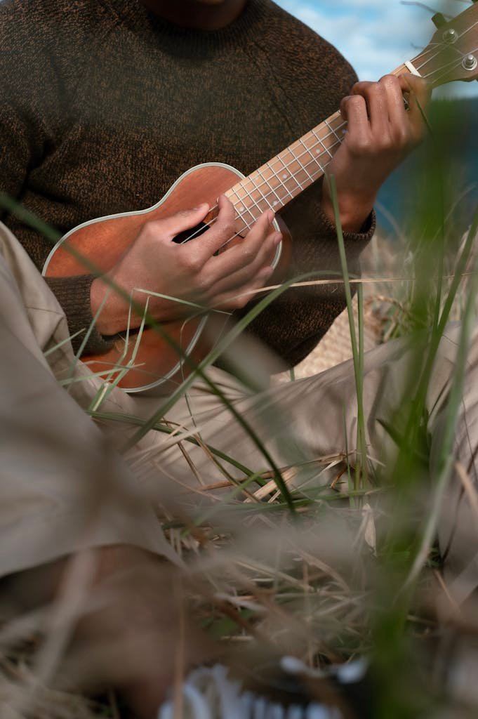 A person strums a ukulele in an outdoor setting surrounded by grass, creating a relaxed atmosphere.